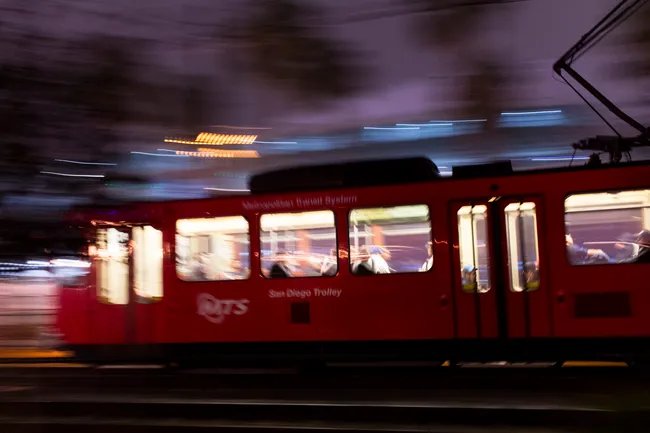 A red San Diego Trolley train moves rapidly at night, captured with motion blur that streaks the background lights into horizontal lines of white and orange. The interior of the trolley is illuminated, revealing blurred figures of passengers through the windows and an open door near the center.