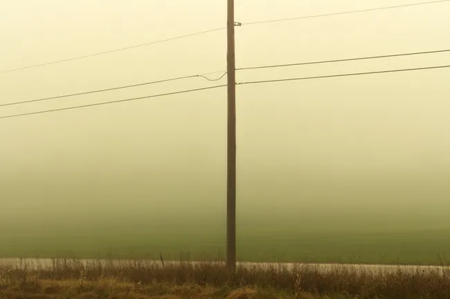 A utility pole stands in the center of a misty, rural landscape. The sky is a hazy, pale yellow-green, fading into a darker green field. In the foreground, there’s a strip of dry, brown grass and a thin, light-colored line of a road. Three power lines stretch horizontally across the upper half of the image from the pole.